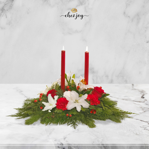 Elegant Christmas candle centerpiece featuring red and white lilies, roses, and carnations, accented with evergreen foliage and red berries. Perfect for holiday décor or gifting, available at Chez Joy Florist.